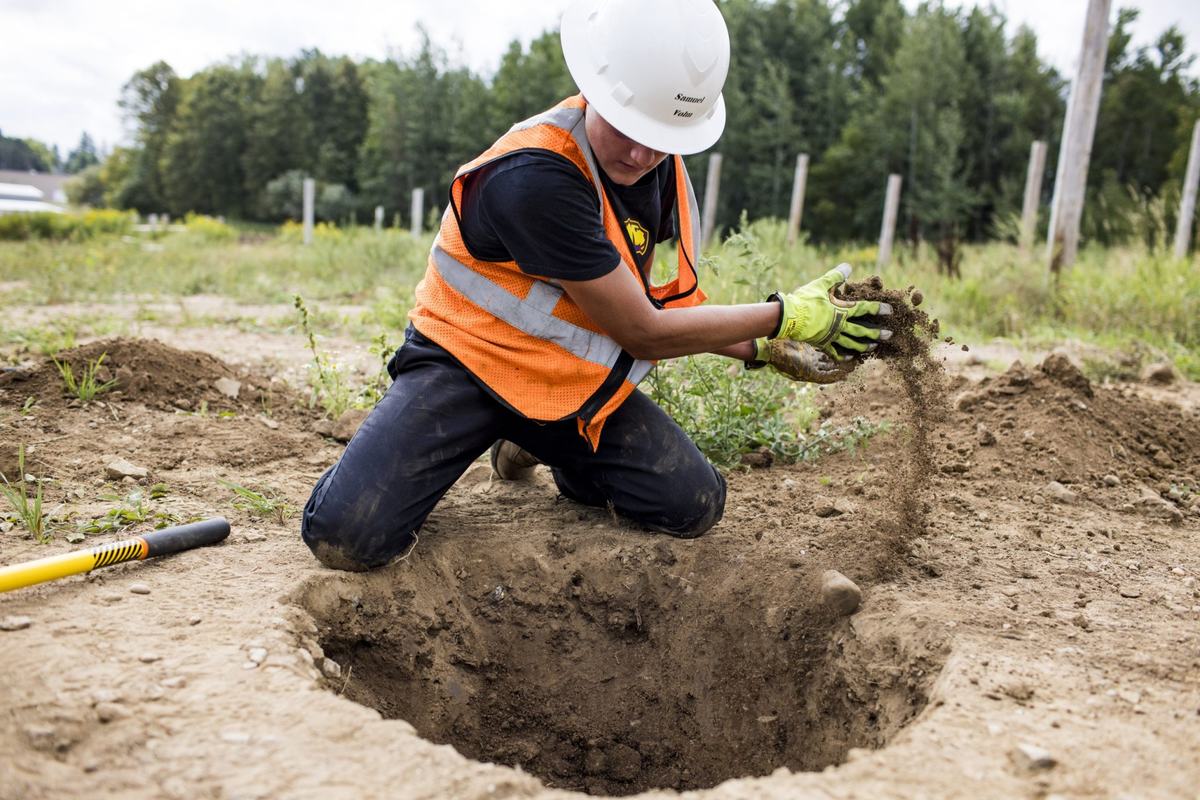 A person wearing a white hard hat, orange reflective vest and yellow gloves kneels beside a large hole in the ground, dropping handfuls of dirt into it.