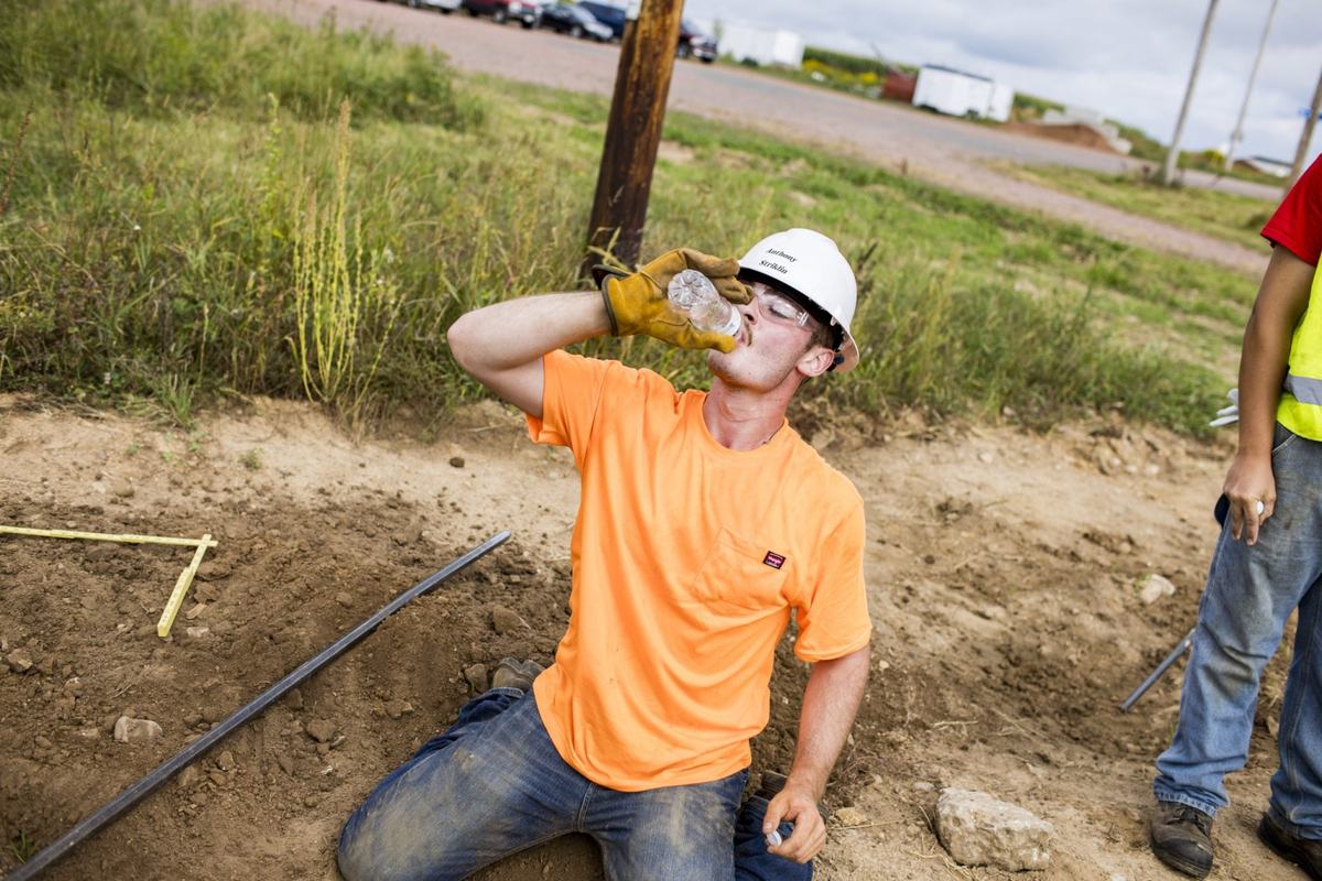 A person wearing a white hard hat, safety glasses, gloves and an orange shirt kneels in a dirt trench while drinking from a clear plastic water bottle.