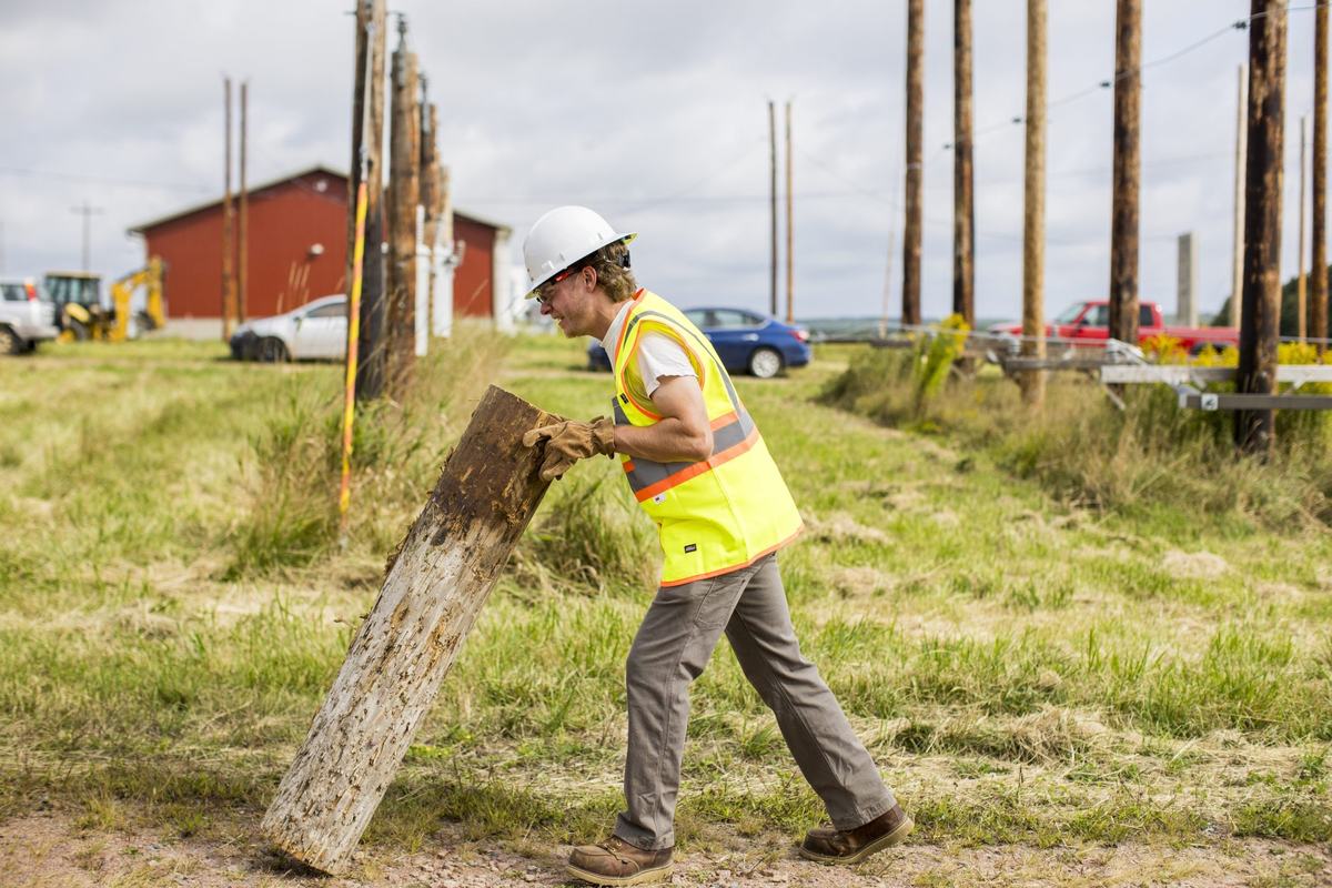 A person wearing a white hard hat, reflective safety vest and gloves holds a large wooden post at an angle in a grassy field lined with utility poles.