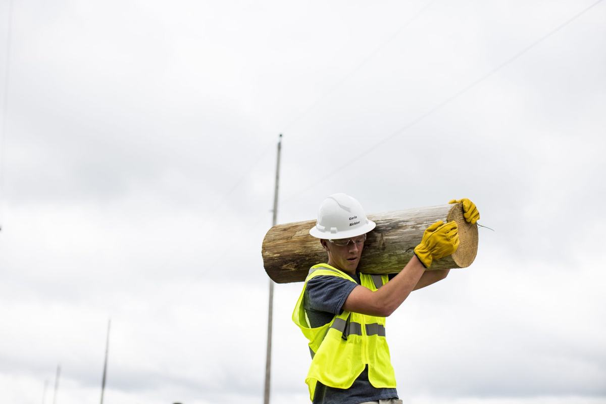 A person wearing a white hard hat, safety glasses, yellow gloves and a reflective vest carries a large chunk of a wooden utility pole on a shoulder beneath a cloudy sky.