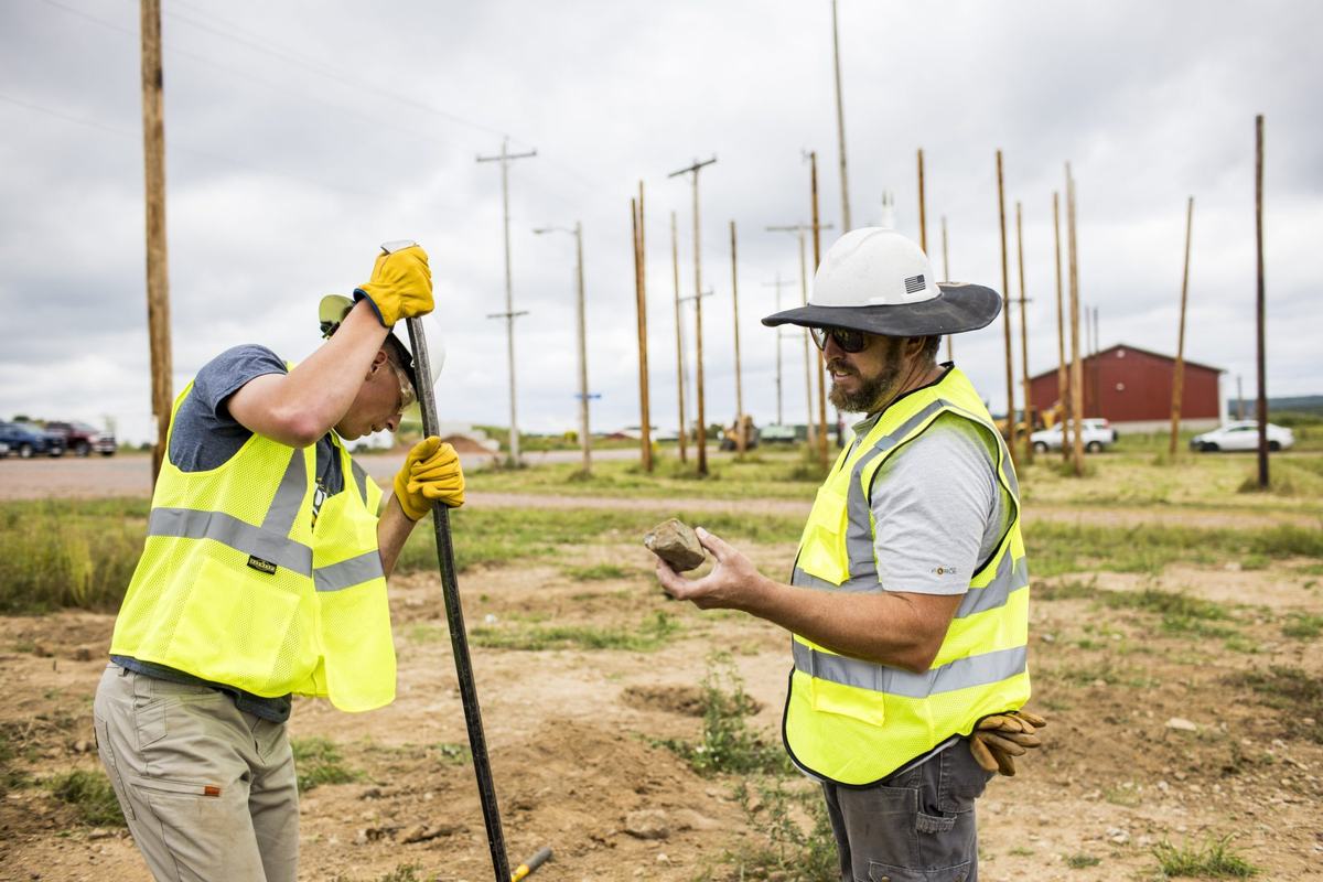 Two people wearing hard hats and reflective safety vests stand in a dirt field. One grips a metal rod while the other holds a rock, with wooden utility poles behind them.
