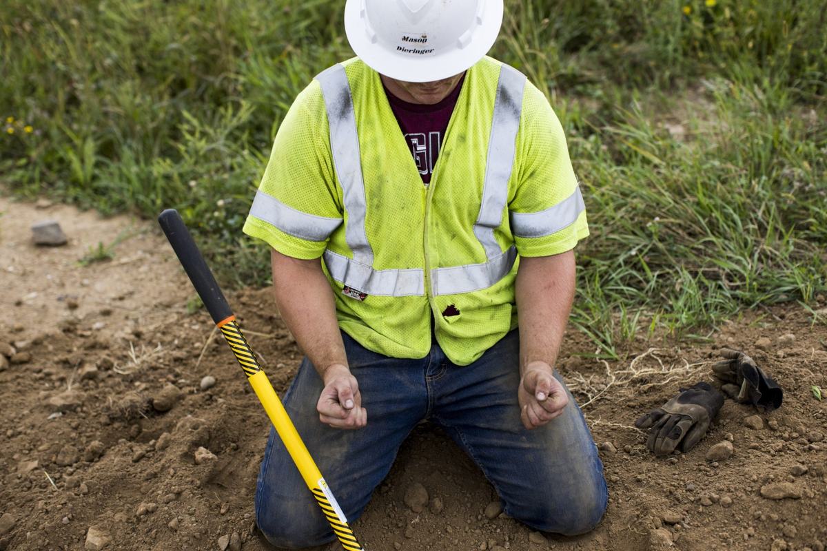 A person wearing a white hard hat and neon yellow reflective vest kneels in dirt, clenching both hands, with a shovel and gloves nearby and grasses in the background.