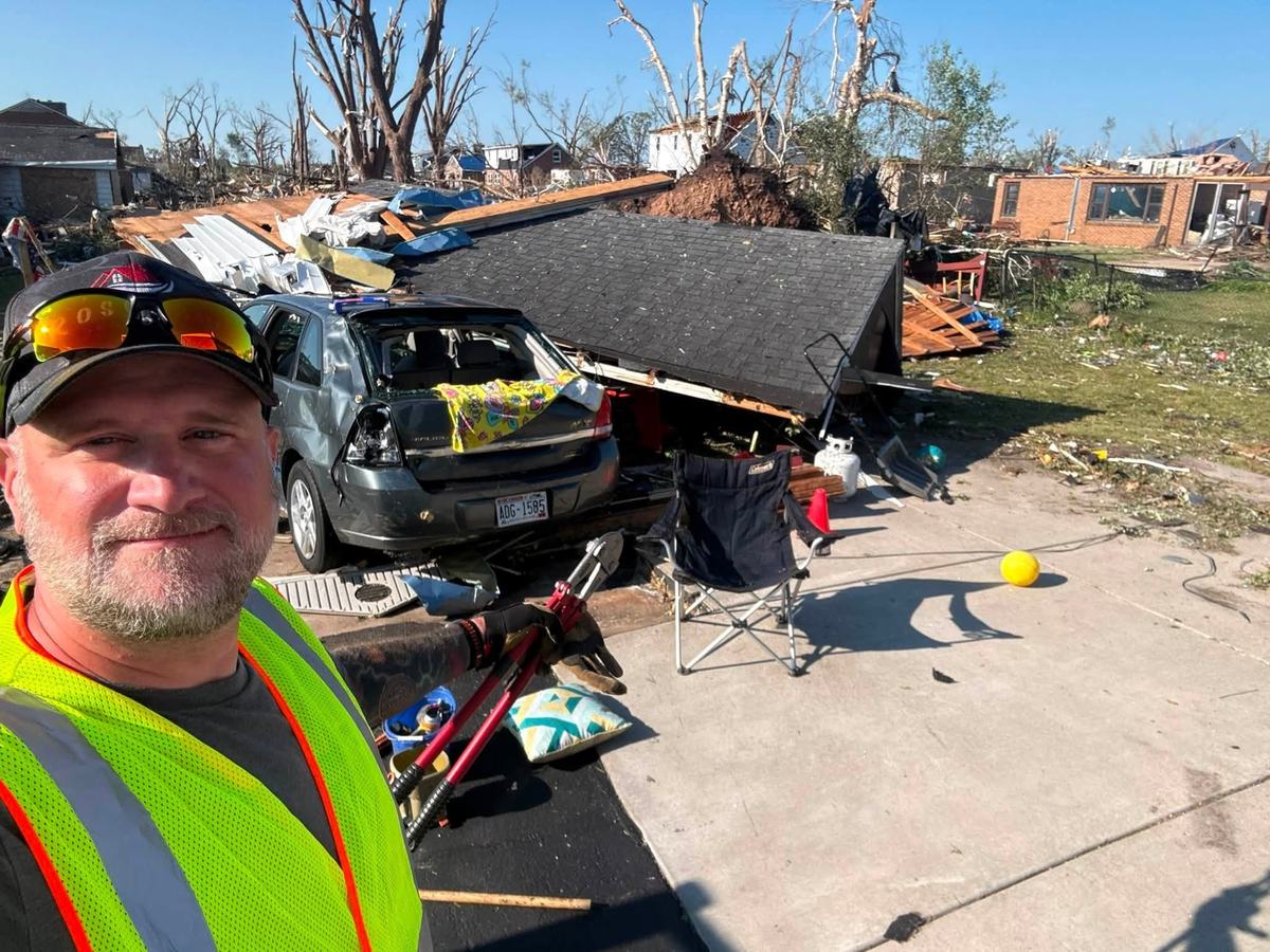 A person in a yellow reflective vest with sunglasses on top of a cap stands near a damaged car and collapsed structure amid broken trees, debris and damaged buildings.