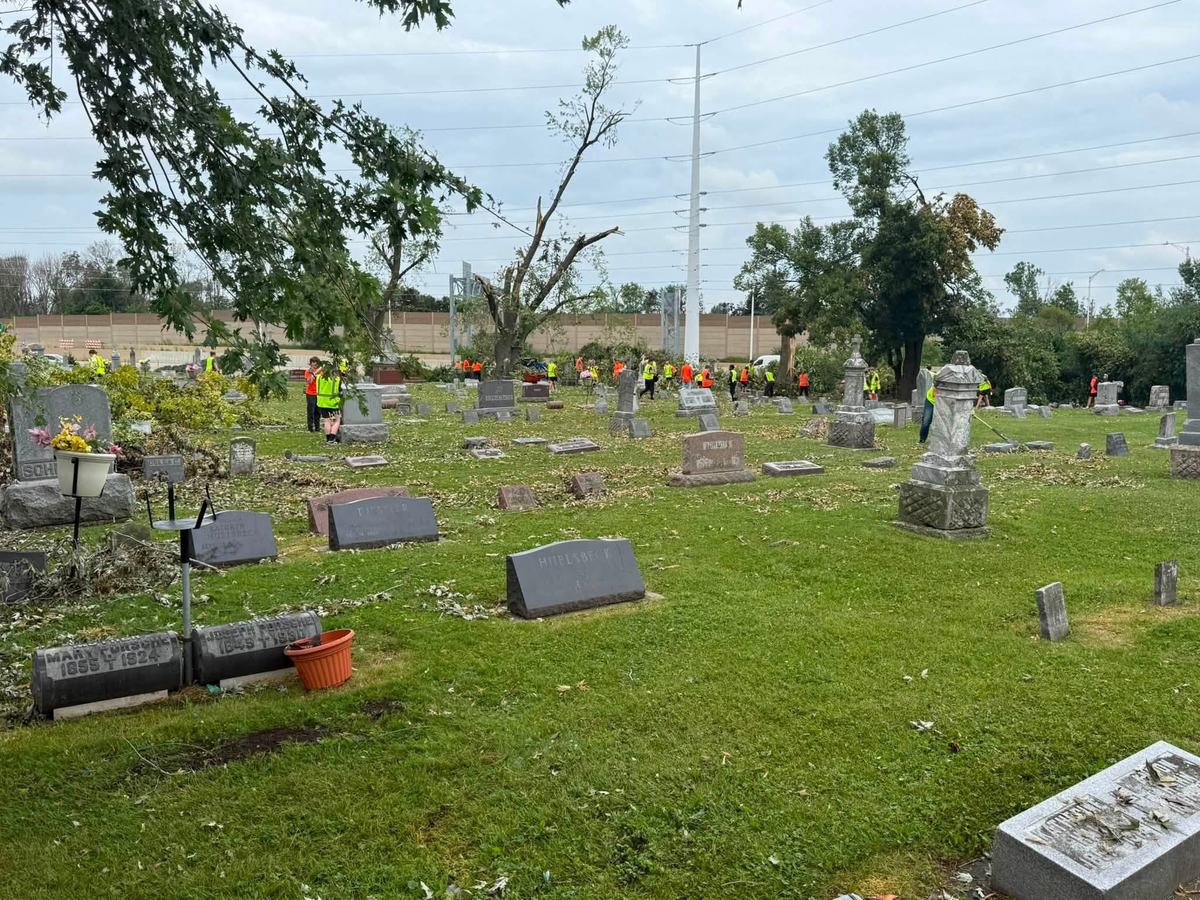 People in bright safety vests stand among gravestones and fallen branches in a cemetery with damaged trees.