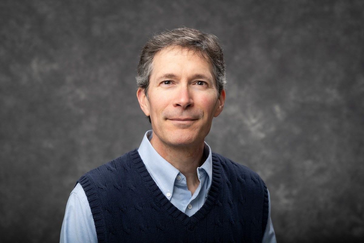 A person with short gray hair wears a light blue collared shirt and dark blue sweater vest against a gray background.