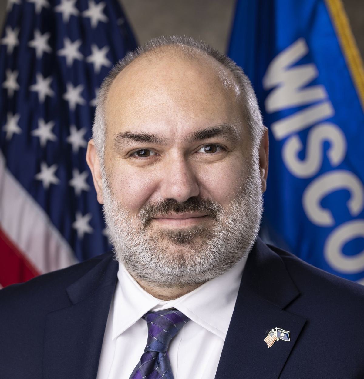 A person with a gray beard wears a dark suit and purple patterned tie, with U.S. and Wisconsin flags in the background.