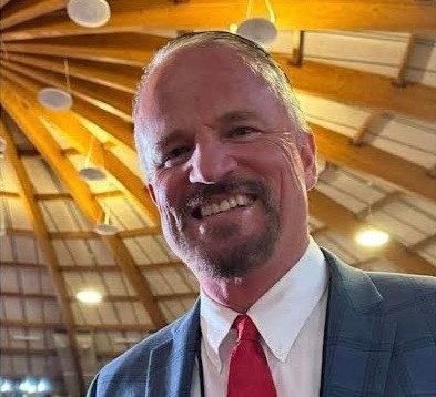A person in a blue plaid suit and red tie smiles at the camera inside a building with exposed wooden roof beams.