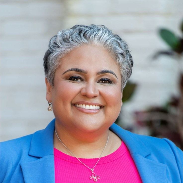 A person with short gray hair wears a bright blue jacket over a pink shirt, a necklace and earrings while smiling at the camera.