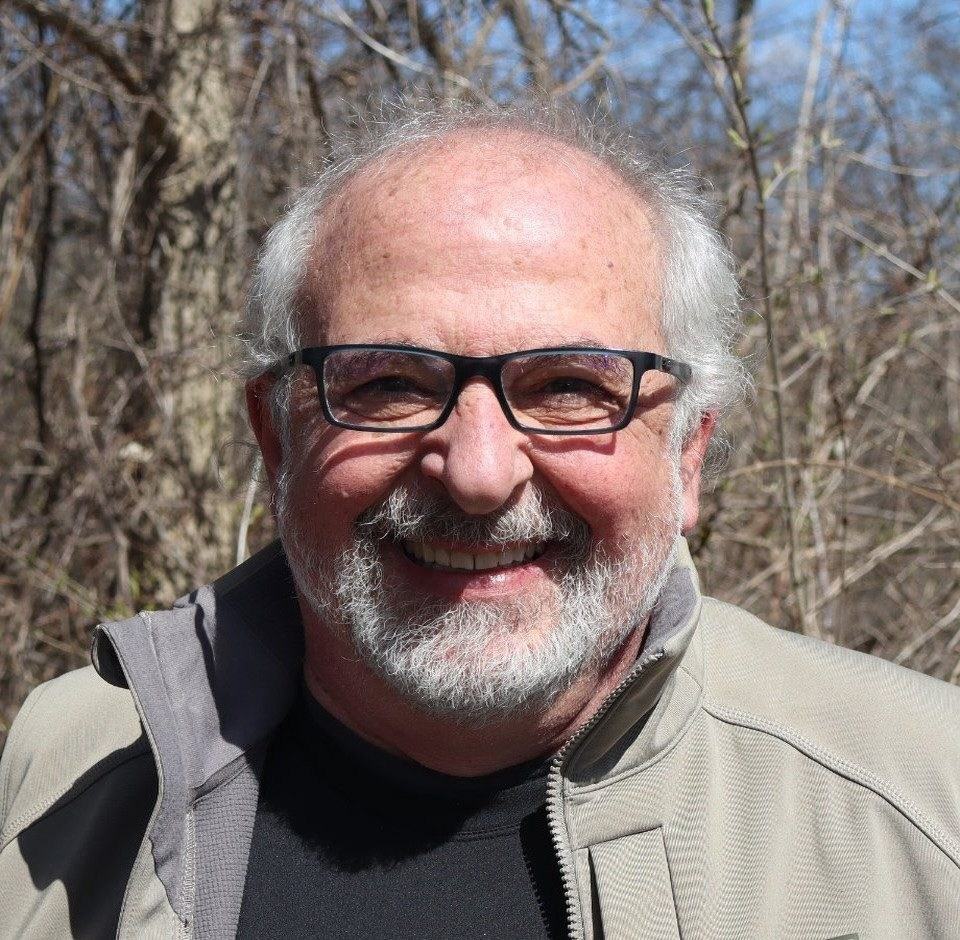 A person with glasses and a white beard smiles at the camera while wearing a gray jacket and black shirt outdoors among leafless trees.