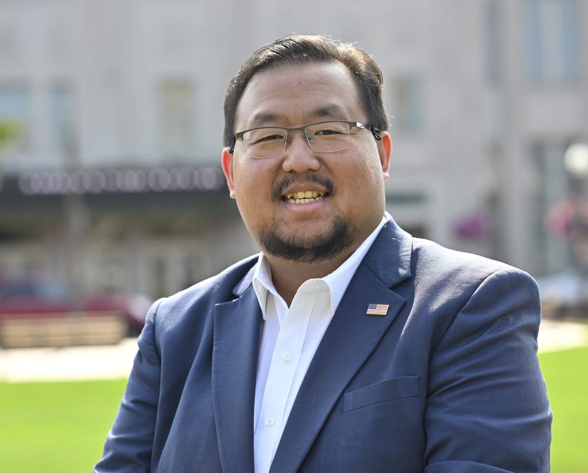 A person with glasses wears a blue suit jacket, white shirt and U.S. flag lapel pin while smiling outdoors.