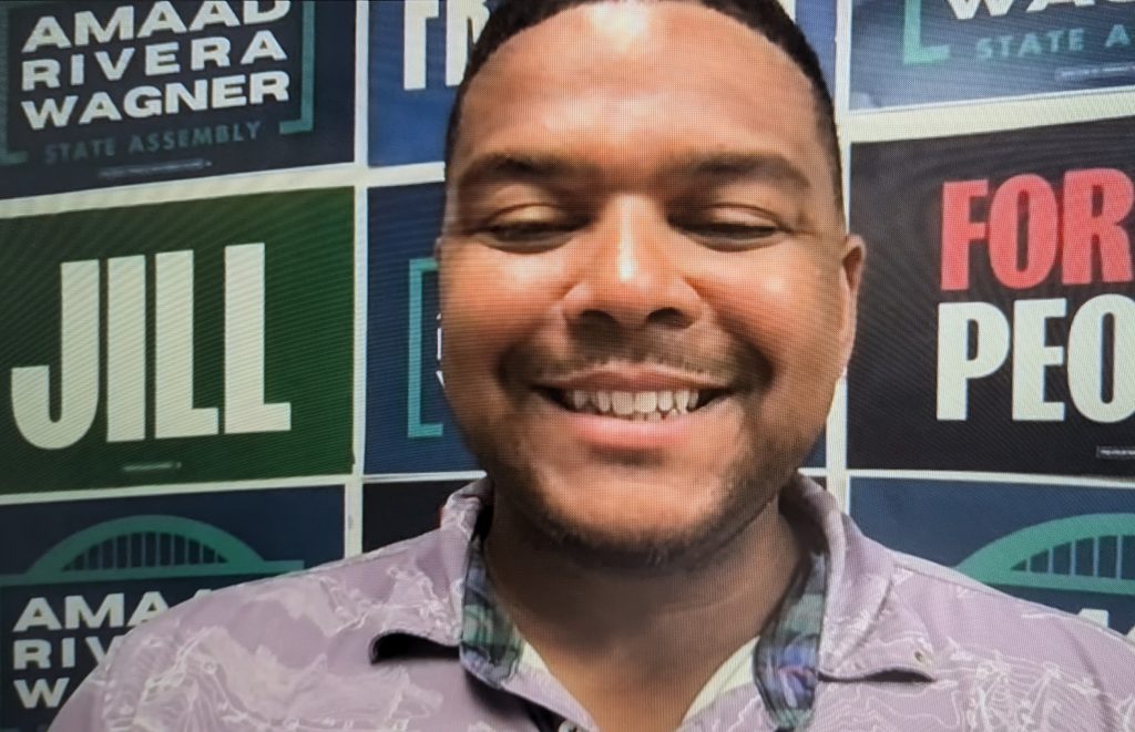 Amaad Rivera-Wagner smiles in front of a wall of political signs for Northeast Wisconsin Democrats.