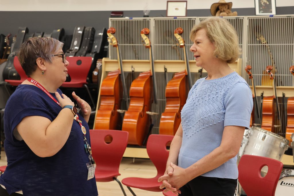 Two people have a conversation inside a ring of chairs, inside a school music room.