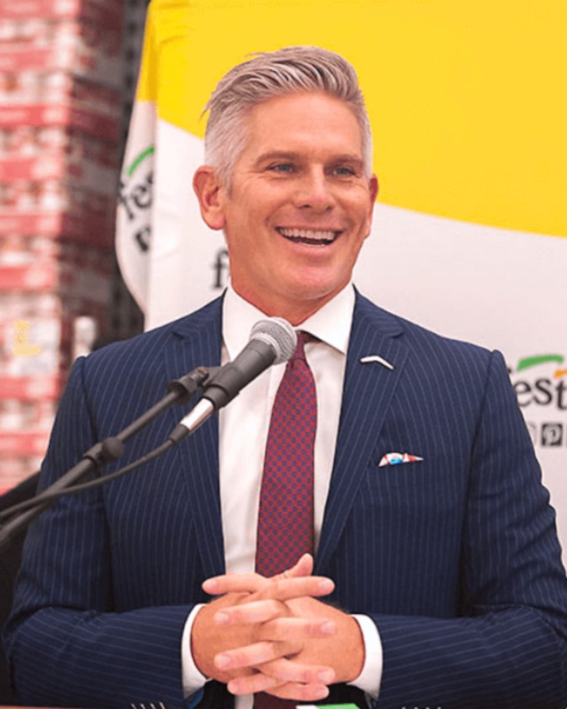 Festival Foods CEO Mark Skogen with hands clasped in front of a Festival Foods banner.
