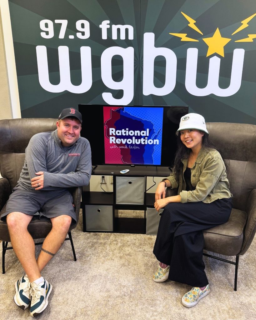 Photo of Rational Revolution Host Mark Becker sitting in a chair next to a TV with the Rational Revolution Logo on it next to Representative Francesca Hong who is also sitting in a chair in the WGBW studio