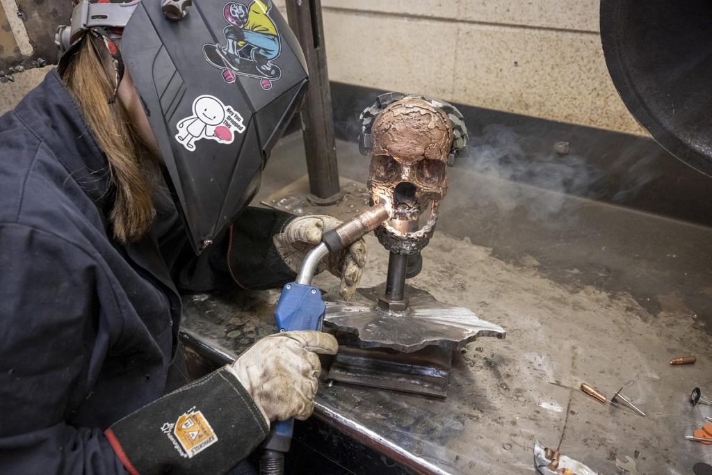 A person wearing gloves and a welding helmet holds a torch next to a metal skull mounted on a stand on a workbench, with smoke rising.