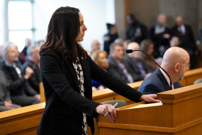 A person stands at a wooden podium holding a pen while another person sits beside a microphone, with rows of seated people blurred in the background.