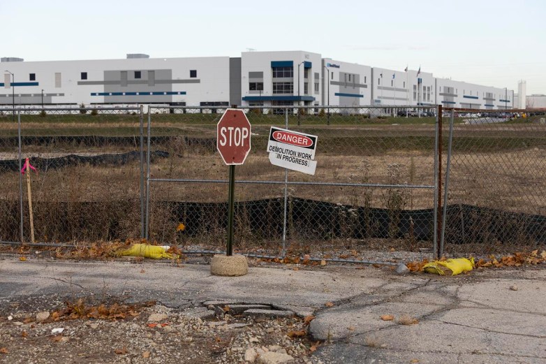 A chain-link fence, a “STOP” sign and a tilted “DANGER Demolition Work in Progress” sign stand in front of an open lot with a large industrial building in the background.