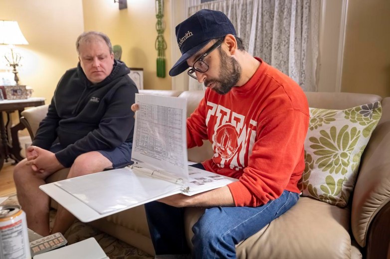 A person in a red sweatshirt and cap sits on a couch examining pages in an open binder while another person sits nearby watching.