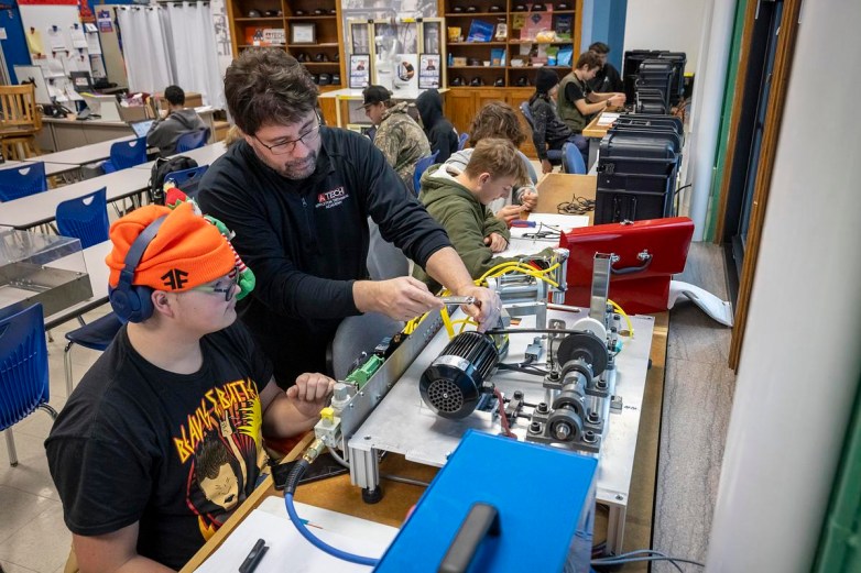 A person points to wiring on a tabletop machine while another person with an orange hat watches while sitting, with several other people working at desks and computers in the background.