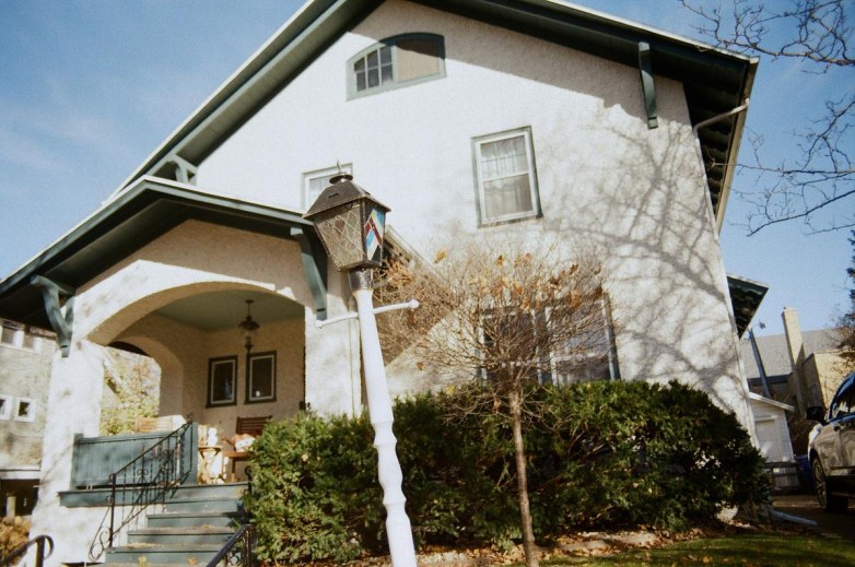 A light-colored house with green trim features an arched front porch, steps with a metal railing, a small tree and bushes, and a decorative lamp post in the yard.