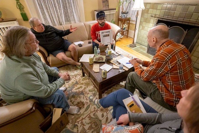 People sit in a room around a coffee table with papers, drinks and snacks as one person holds up a printed page. A fireplace, a lamp, a plant and other items are in the room.