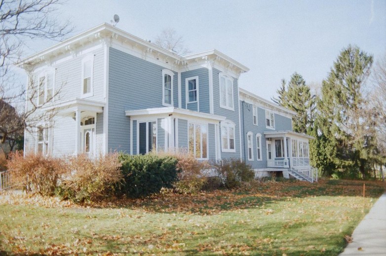 A large blue house with white trim and multiple tall windows, a small porch, and surrounding shrubs and trees with fallen autumn leaves on the lawn.