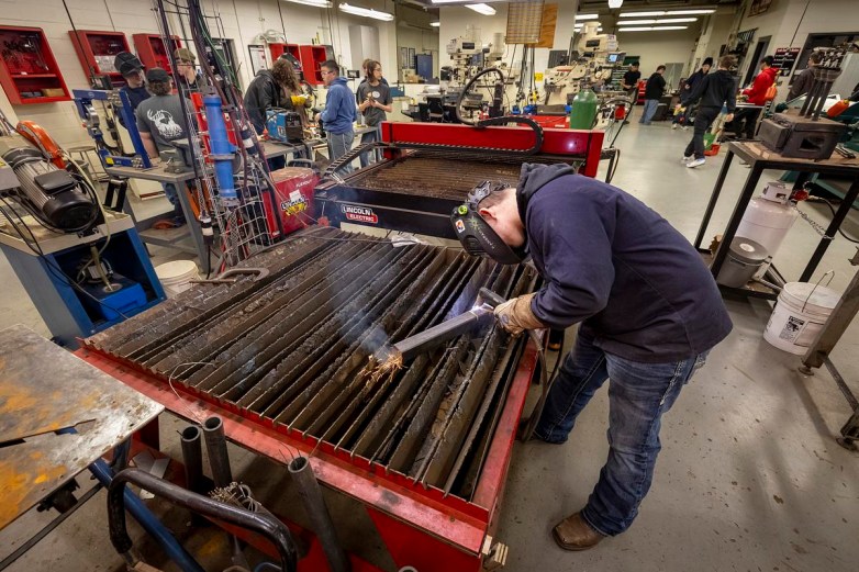 A person wearing gloves and a welding helmet uses a torch on a metal sheet atop a large table, with sparks flying and several other people and machines visible in a workshop.