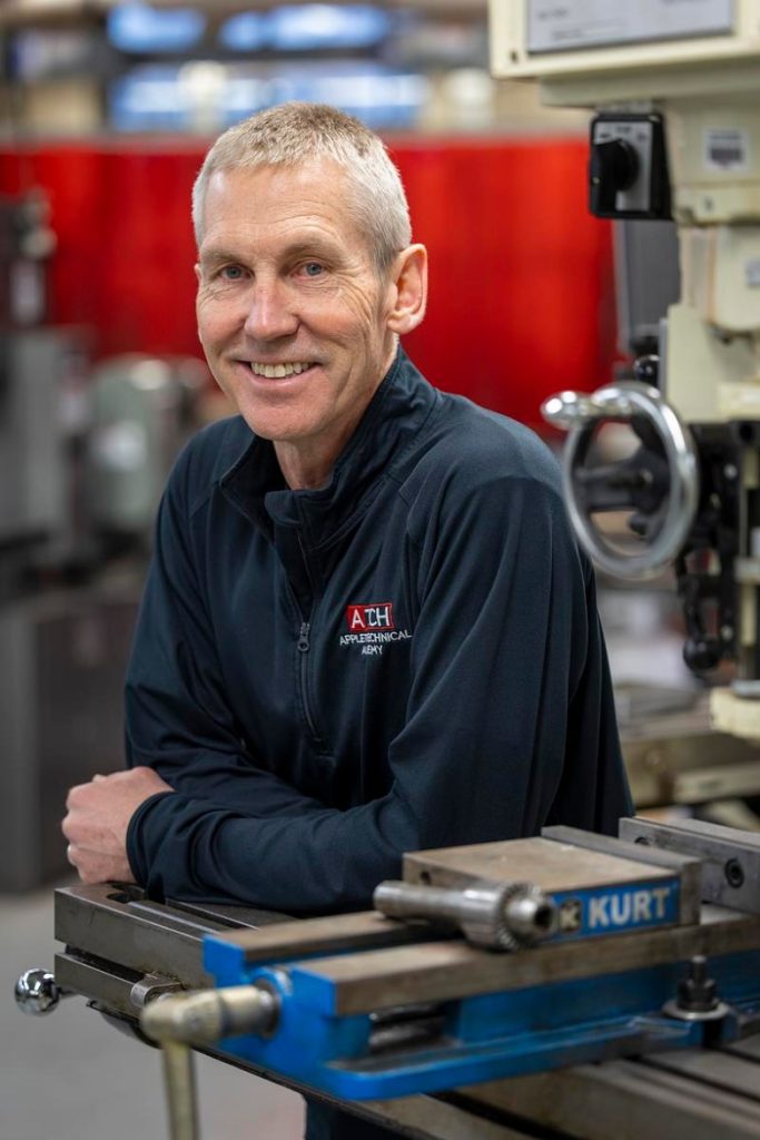 A person smiles and leans on a metal machine table in a workshop, wearing a dark top with a logo reading “Atech Appleton Technical Academy,” with industrial equipment in the background.