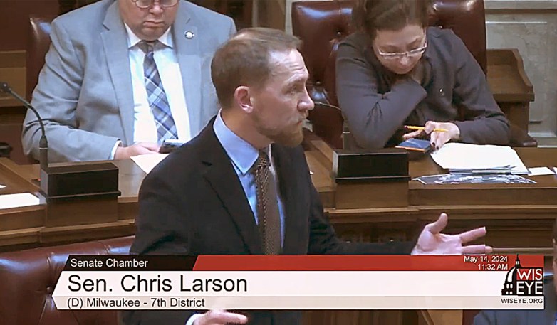 A person gestures with one hand while others sit behind at desks as on-screen text reads “Sen. Chris Larson”