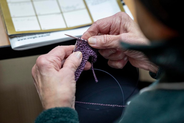 A person’s hands hold knitting needles and purple yarn, forming small stitches over a table with papers nearby.