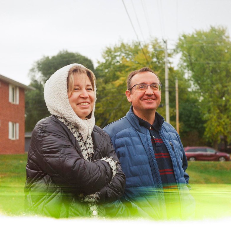 Two people stand outdoors wearing jackets, smiling and looking ahead, with trees, a grassy area and a brick building visible in the background.