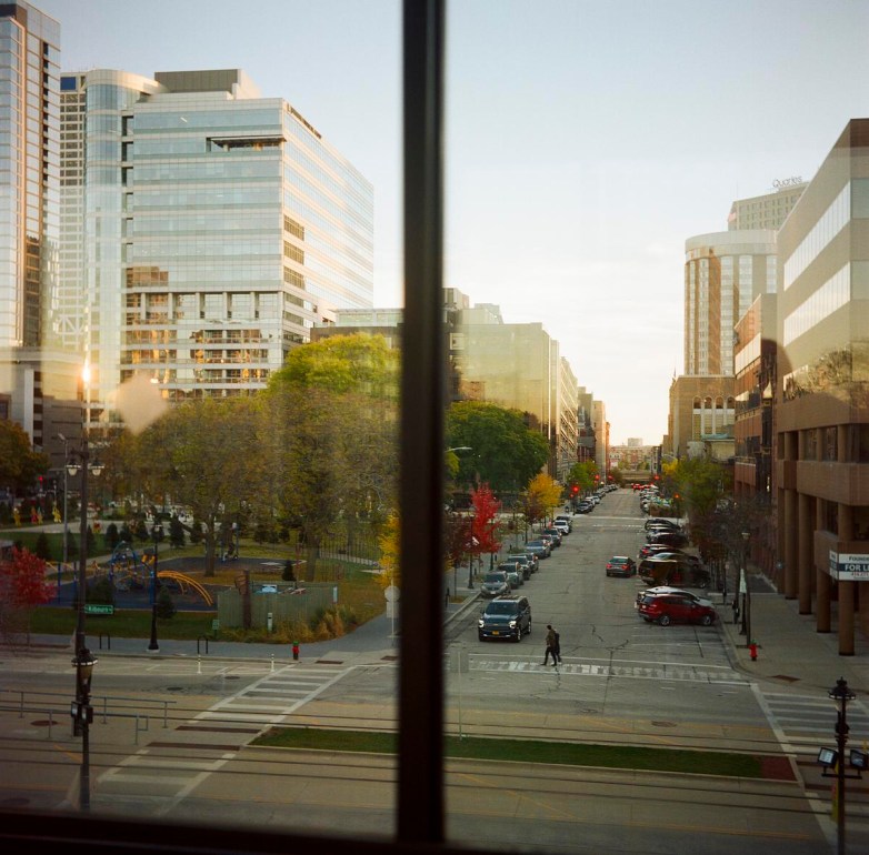 A city street lined with buildings, trees and parked cars is seen through a window, with a crosswalk and pedestrians visible below and glass office buildings in the distance.