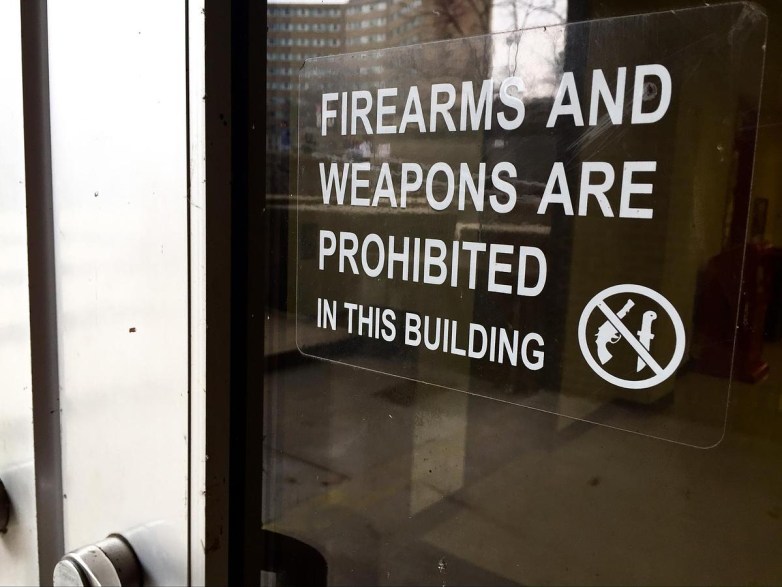 A sign on a glass door reads “FIREARMS AND WEAPONS ARE PROHIBITED IN THIS BUILDING,” with a crossed-out gun icon beside the text.