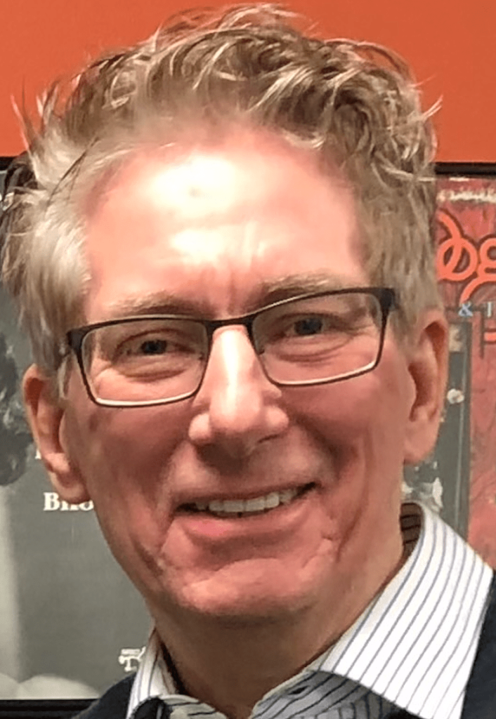 A person wearing glasses smiles slightly in a close-up portrait, with short hair and a framed poster on a wall in the background.