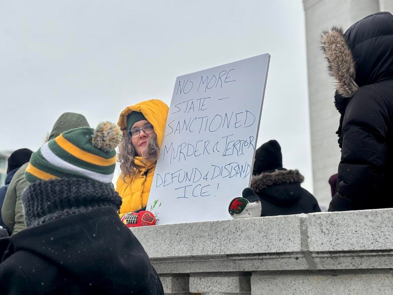 A person in winter clothing holds a handwritten sign reading “NO MORE STATE SANCTIONED MURDER & TERROR DEFUND & DISBAND ICE!” while others stand nearby outdoors.