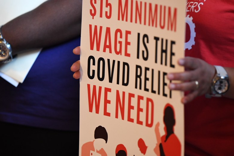 A person holds a sign reading “$15 MINIMUM WAGE IS THE COVID RELIEF WE NEED,” with illustrated figures at the bottom and hands gripping the edges.